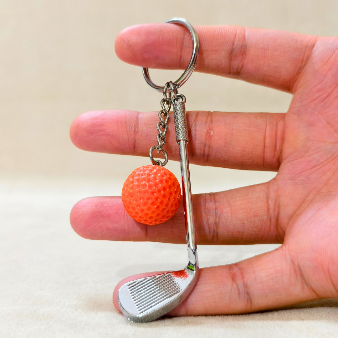 A miniature golf club keychain with an orange textured golf ball charm, held between a person's fingers against a neutral beige background.
