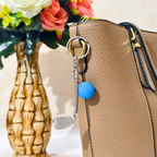 A tan textured leather tote bag with a silver-tone key ring and a golf-themed keychain charm featuring a silver golf club head and a blue textured golf ball, styled next to a decorative vase of flowers.
