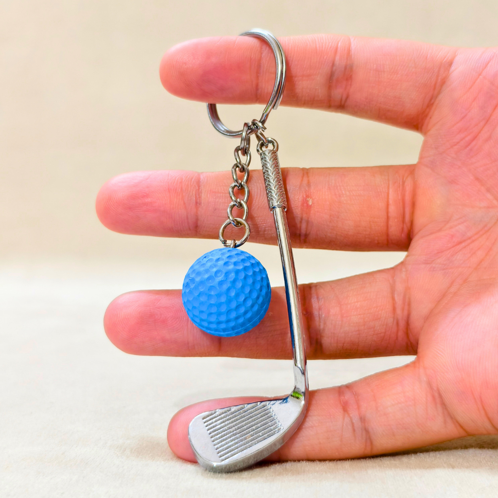 A miniature golf keychain held in a hand, featuring a silver metal golf club head and a small blue textured golf ball charm.