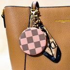 A close-up of a tan pebbled leather handbag with a round pink and brown checkerboard keychain charm and a matching patterned scarf attached by gold hardware.