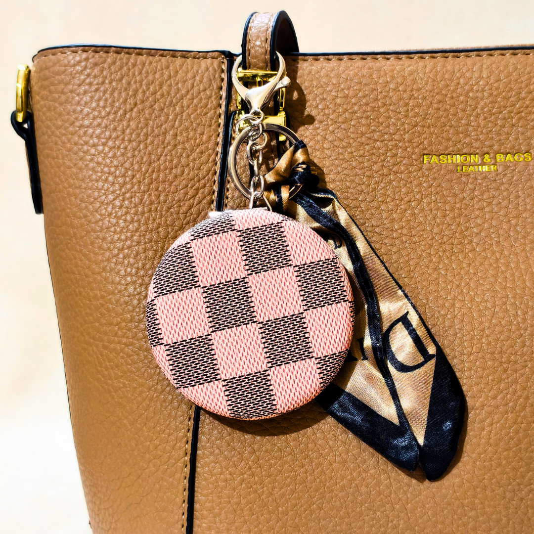 A close-up of a tan pebbled leather handbag with a round pink and brown checkerboard keychain charm and a matching patterned scarf attached by gold hardware.