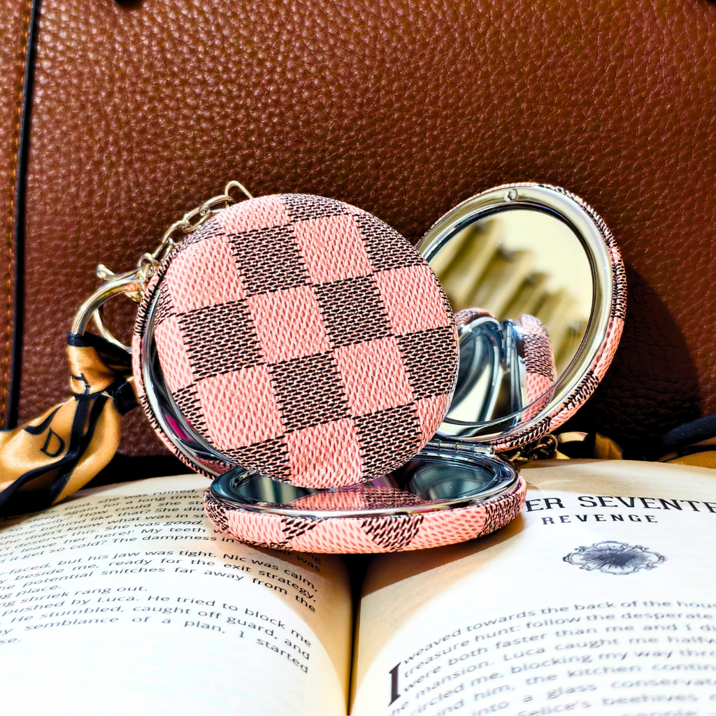 A compact round mirror with a pink and dark brown checkerboard patterned exterior, shown open with a mirror inside, resting on an open book against a brown textured background.