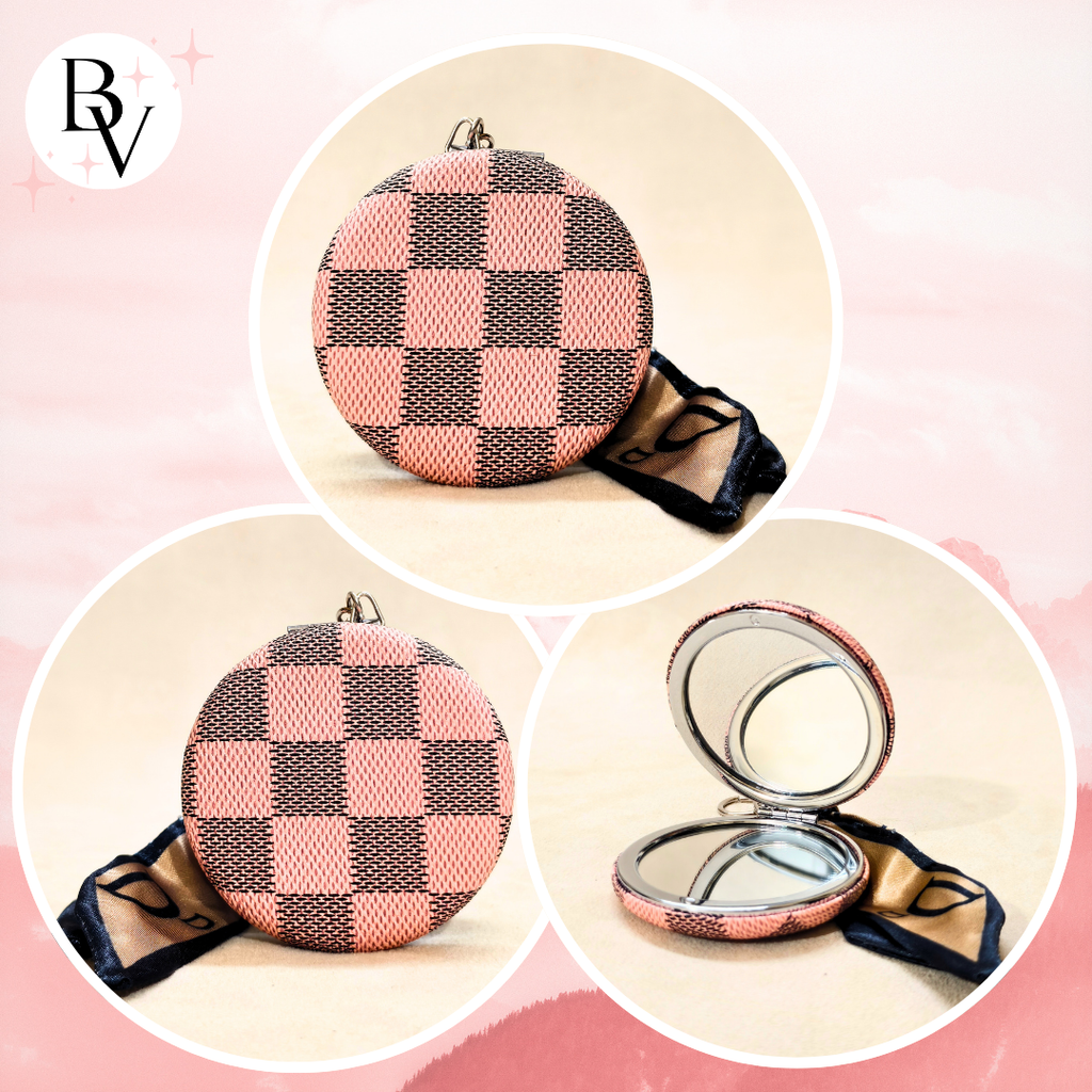 A round compact mirror with a pink and dark brown checkerboard patterned cover, shown in three views: closed, closed with a black ribbon, and open with the mirror side visible. A 'BV' logo is in the top left corner.