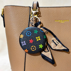 A close-up of a tan pebbled leather handbag with a black circular keychain cover featuring colorful Louis Vuitton-style monogram motifs and a beige-and-black patterned scarf charm attached by a gold clasp.