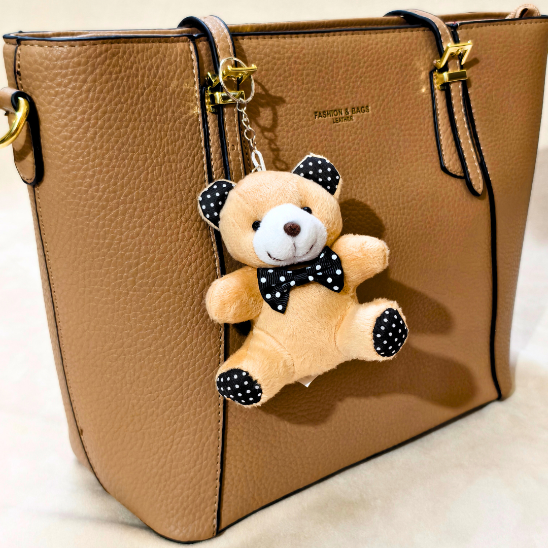 A tan pebbled leather tote bag with gold-tone hardware and a hanging plush teddy bear keychain. The bear is cream-colored with black polka-dot ears, bow tie, and paw pads, shown against a light neutral background.