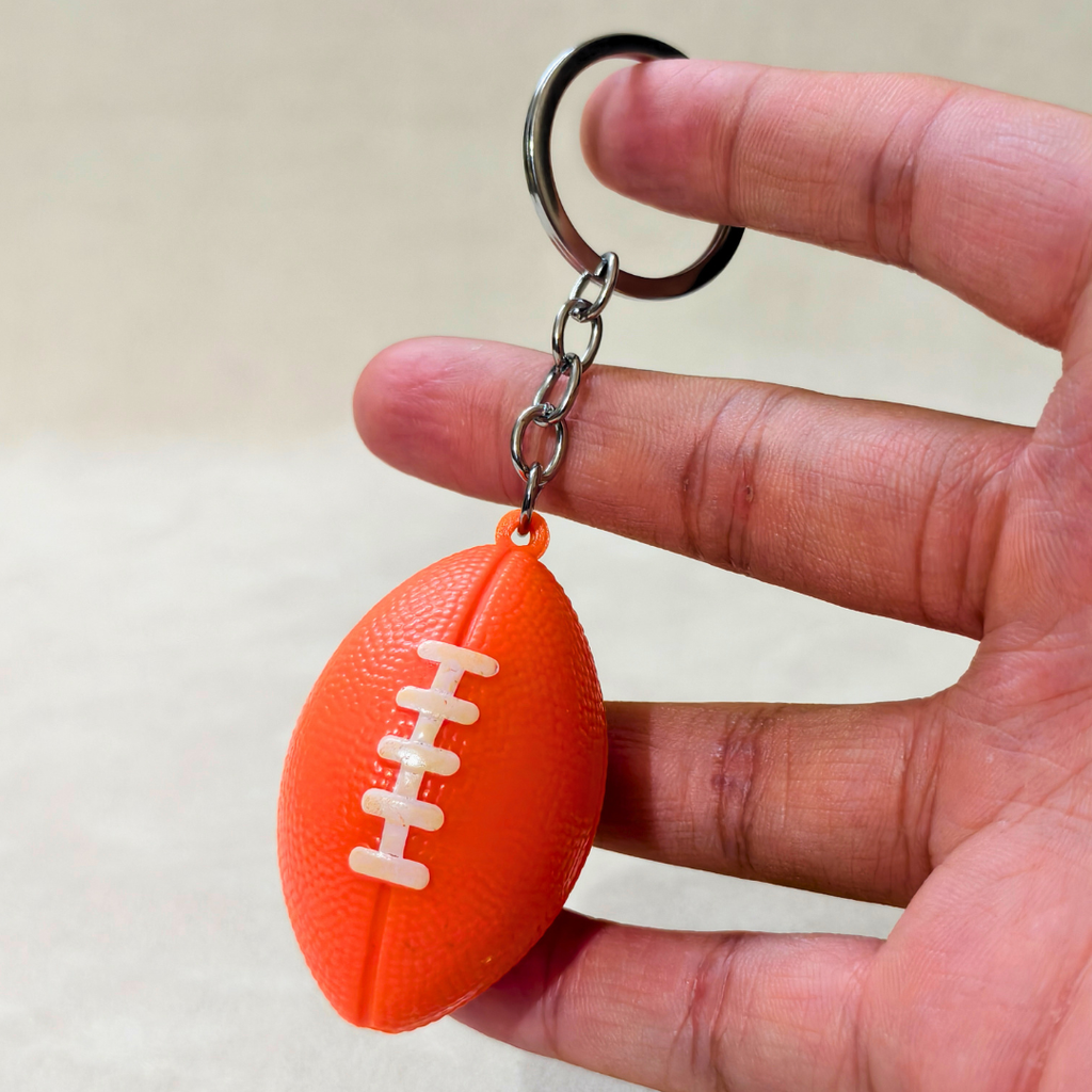An orange football-shaped keychain with white laces and a silver metal key ring, held between fingers against a light beige background.