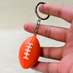 An orange football-shaped keychain with white laces and a silver metal key ring, held between fingers against a light beige background.