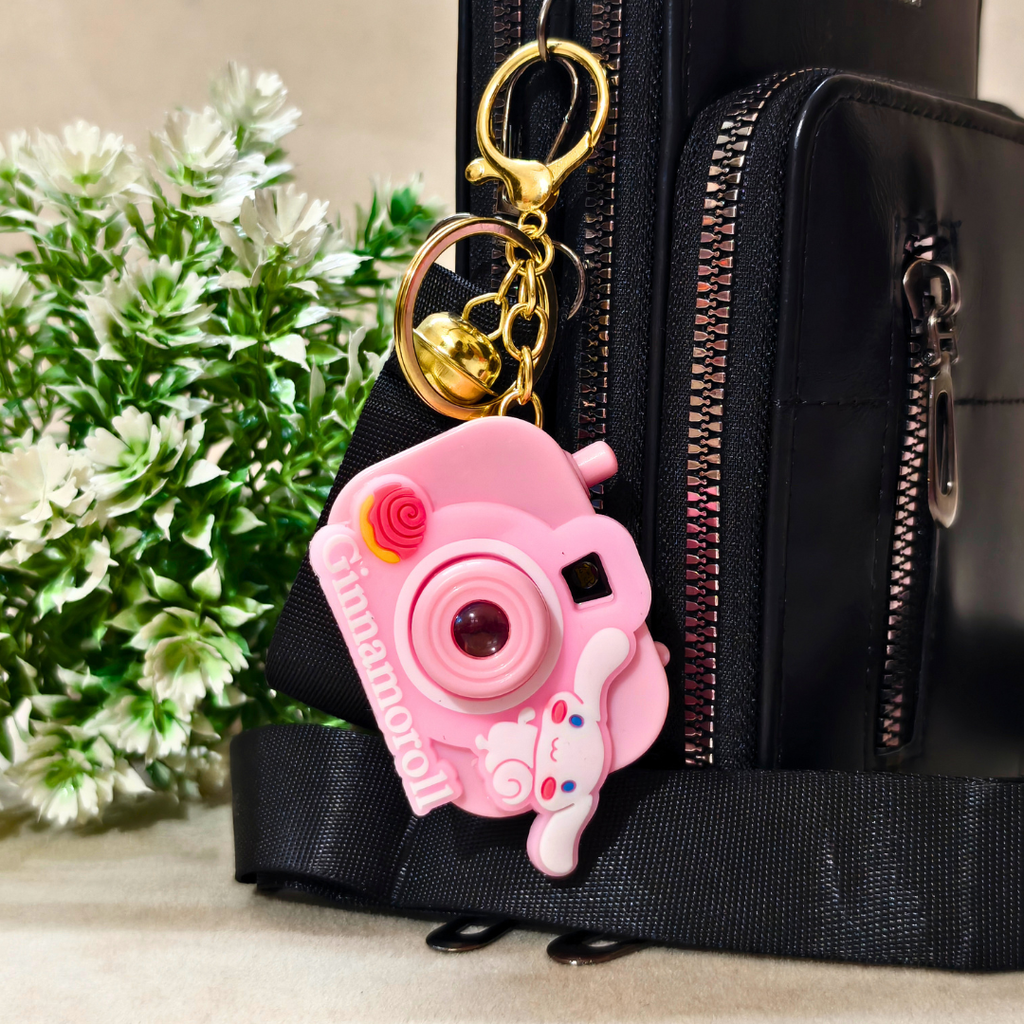 Pink camera-shaped keychain attached to a black bag with a plant in the background