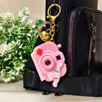 Pink camera-shaped keychain attached to a black bag with a plant in the background