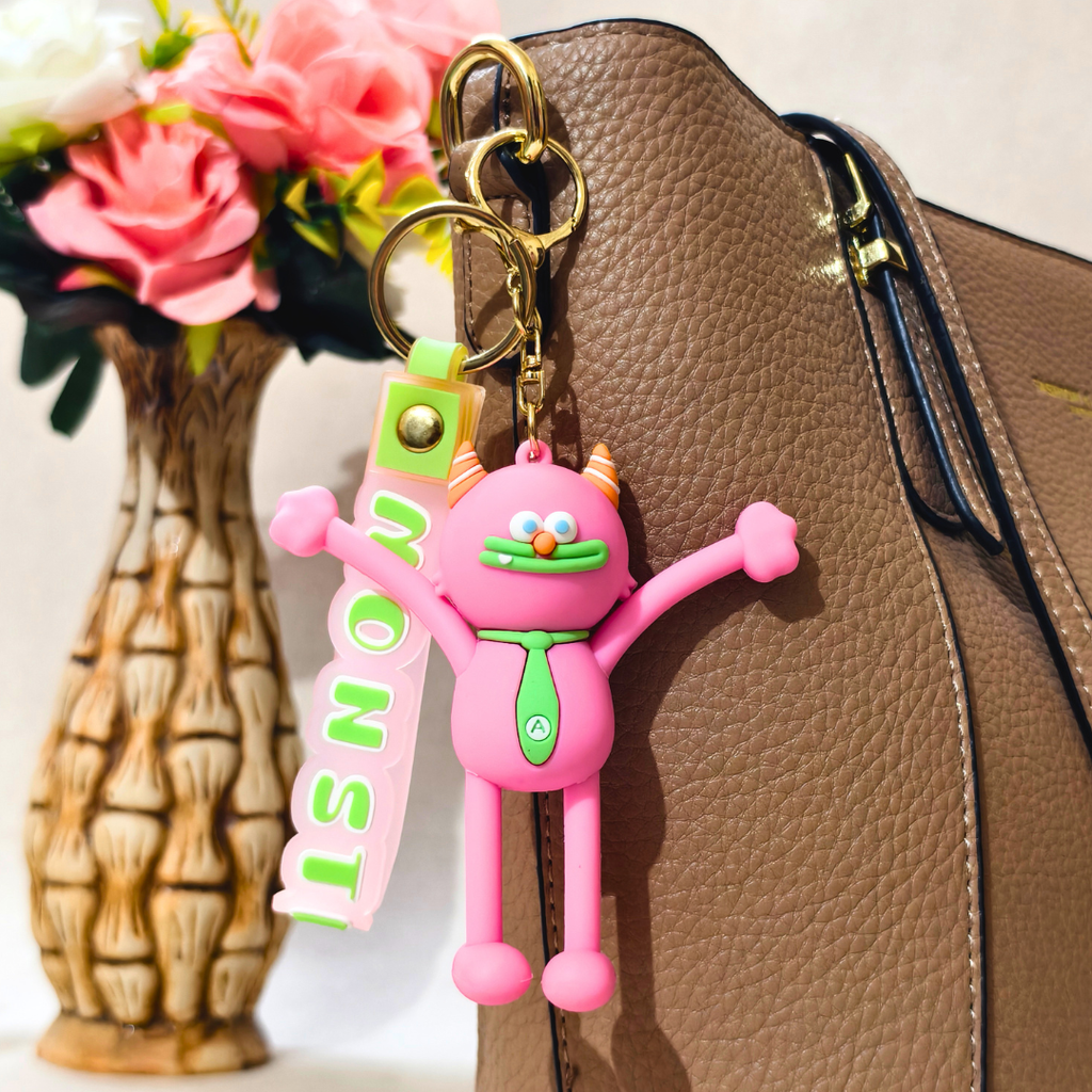 A pink cartoon monster keychain charm with green tie details and a vertical pink strap reading 'MONST', attached to a gold key ring and shown hanging beside a brown textured handbag with flowers in a vase in the background.