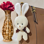 White plush bunny keychain attached to a brown leather bag with a red flower in the background.