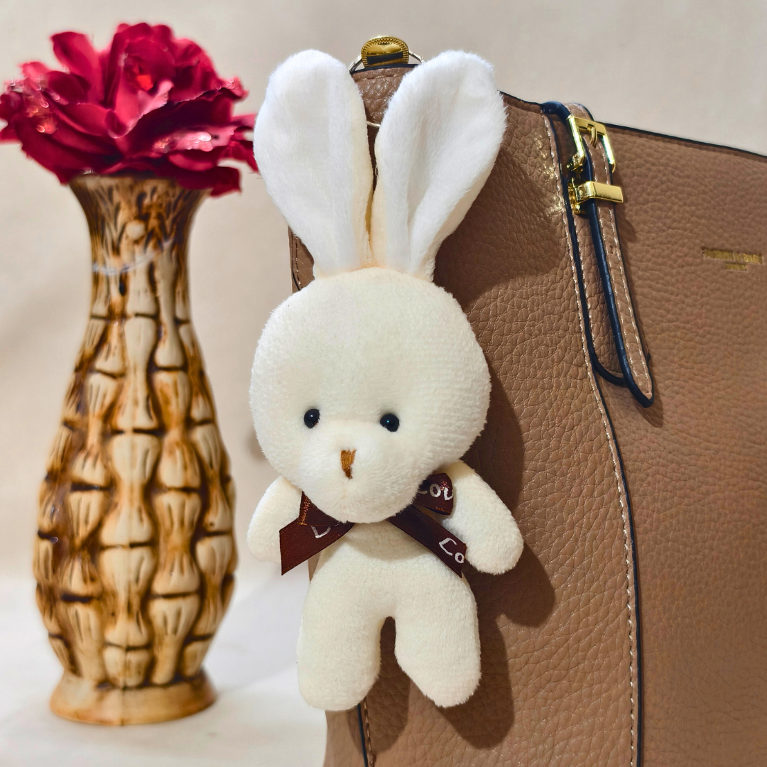 White plush bunny keychain attached to a brown leather bag with a red flower in the background.