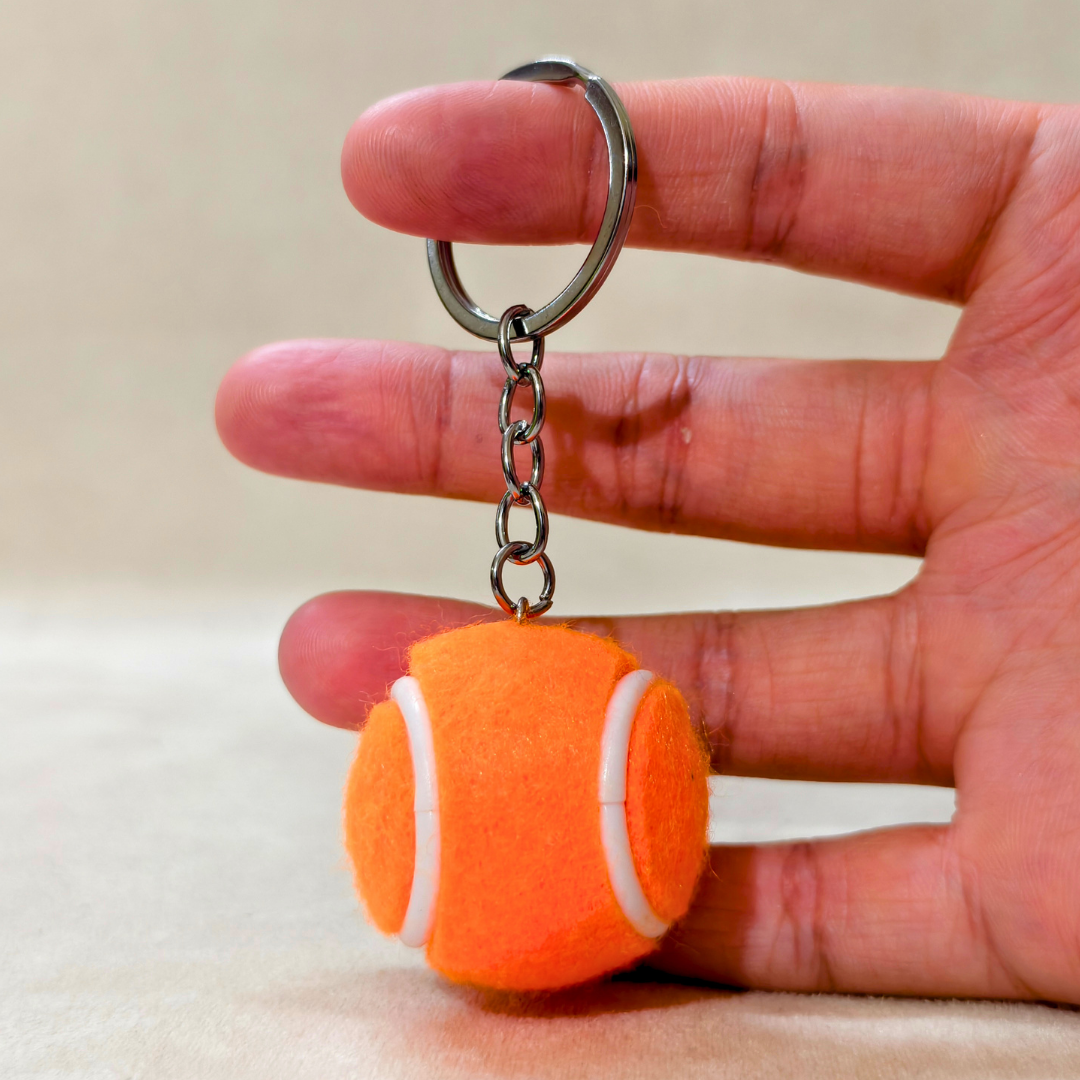An orange tennis ball keychain held between a person's fingers. The keychain features a bright orange felt tennis ball charm with white curved lines, attached to a silver metal chain and split ring.