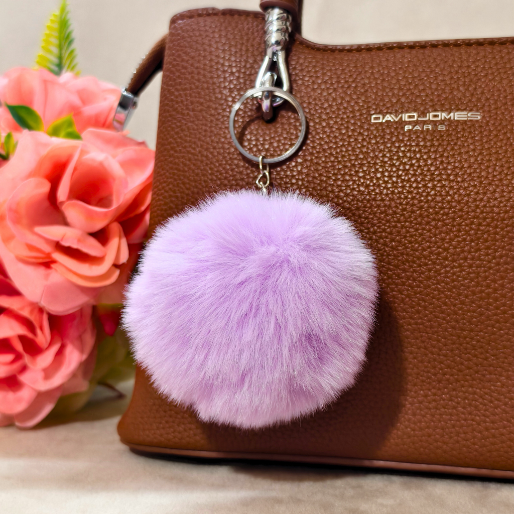 Purple fluffy keychain attached to a brown handbag with pink flowers in the background