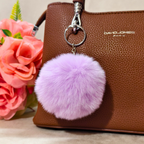 Purple fluffy keychain attached to a brown handbag with pink flowers in the background