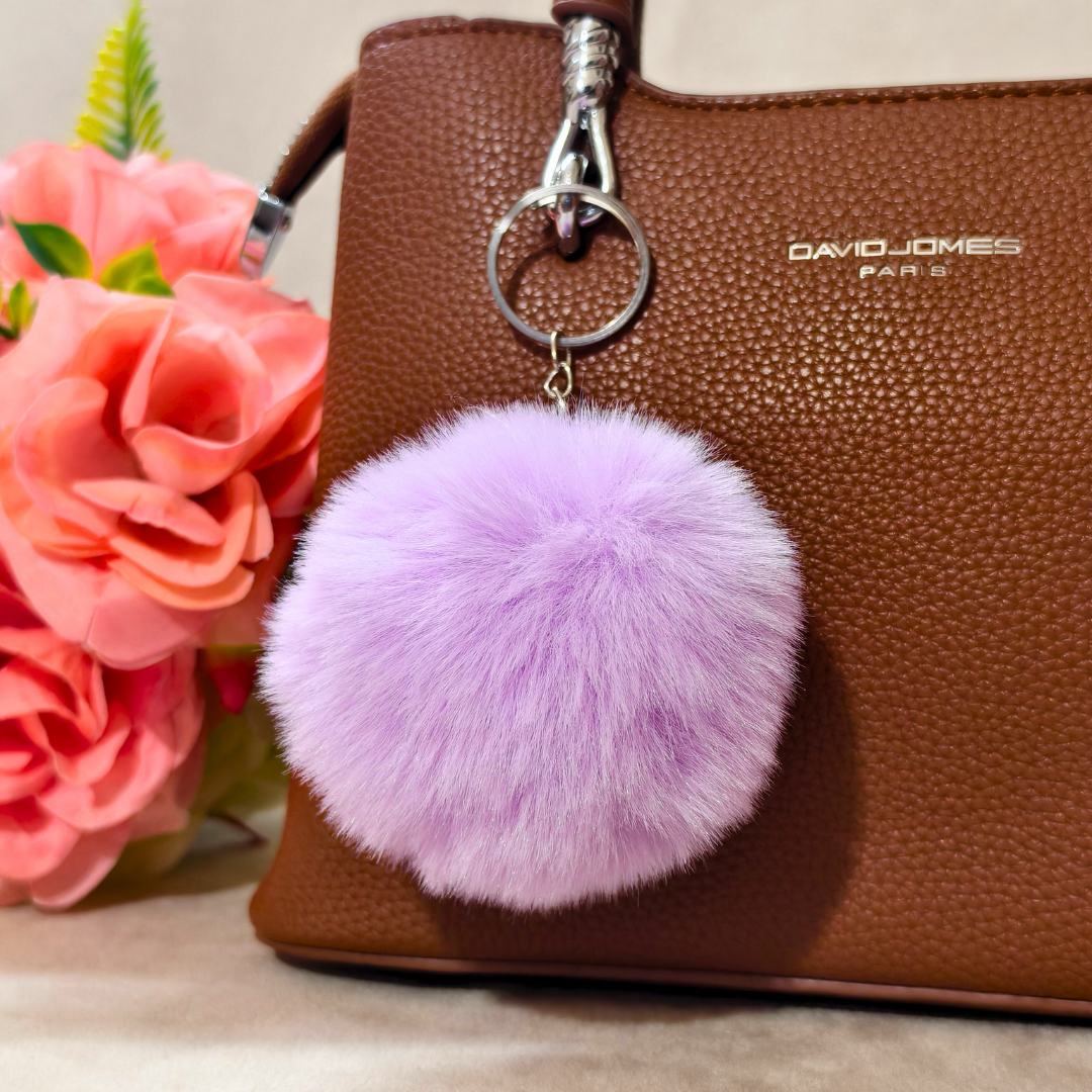 Purple fluffy keychain attached to a brown handbag with pink flowers in the background