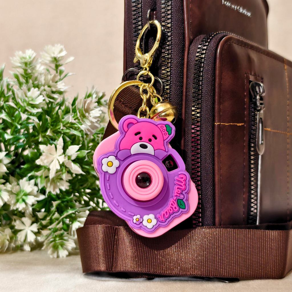 A pink and purple cartoon bear camera keychain charm with a gold clasp, shown attached to a brown leather bag next to white flowers.