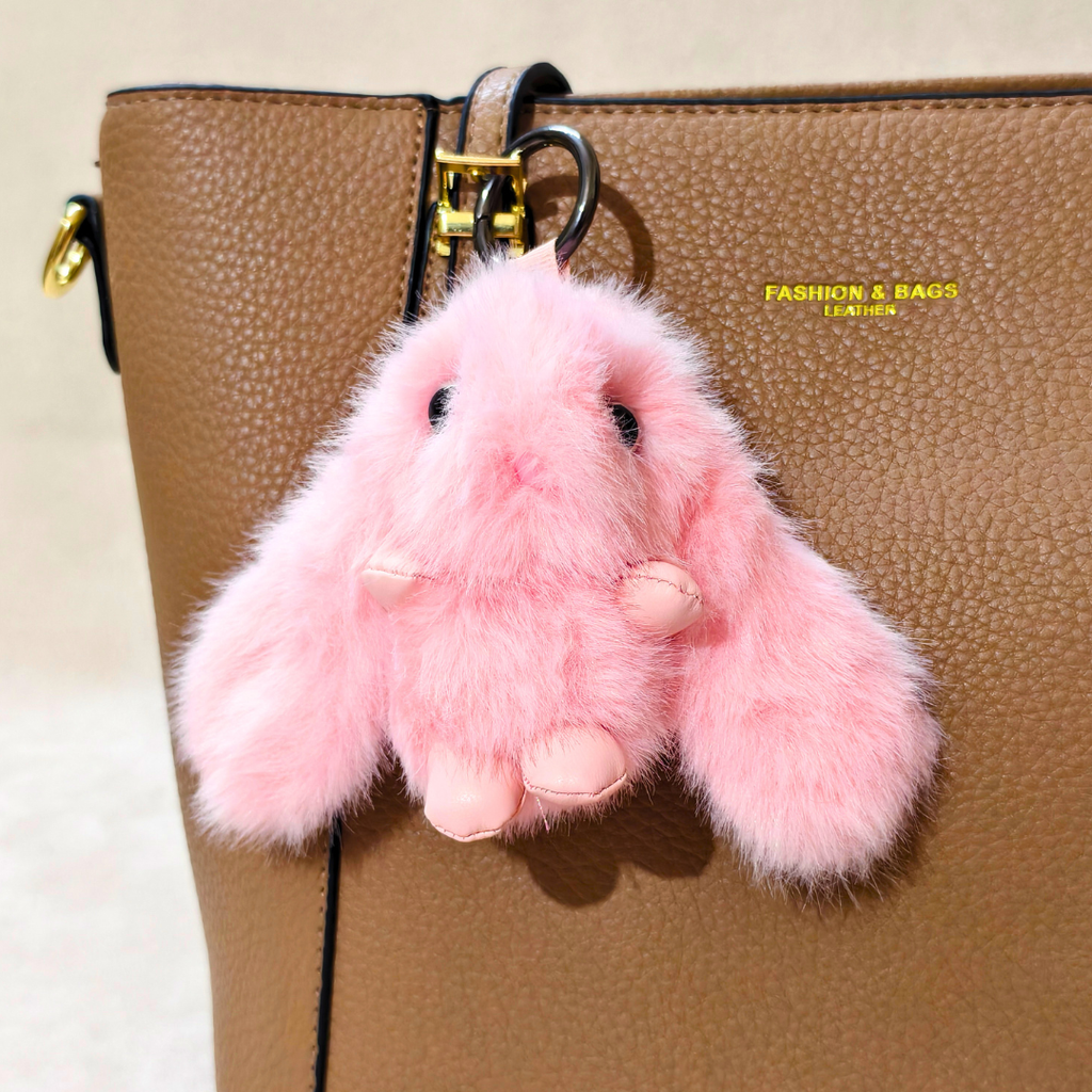 A pink fluffy bunny keychain charm with black embroidered eyes and nose, hanging from a gold ring on a brown textured leather handbag. The bag has yellow text reading 'FASHION BAGS' on the side.