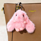 A pink fluffy bunny keychain charm with black embroidered eyes and nose, hanging from a gold ring on a brown textured leather handbag. The bag has yellow text reading 'FASHION BAGS' on the side.