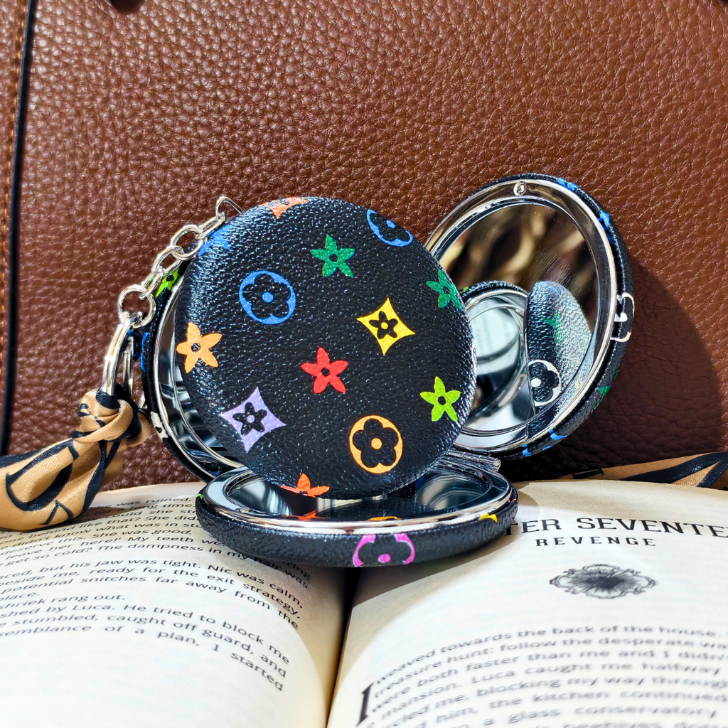 A small round black compact mirror with a colorful monogram-style floral print, shown open on top of an open book against a brown textured leather background.