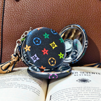 A small round black compact mirror with a colorful monogram-style floral print, shown open on top of an open book against a brown textured leather background.