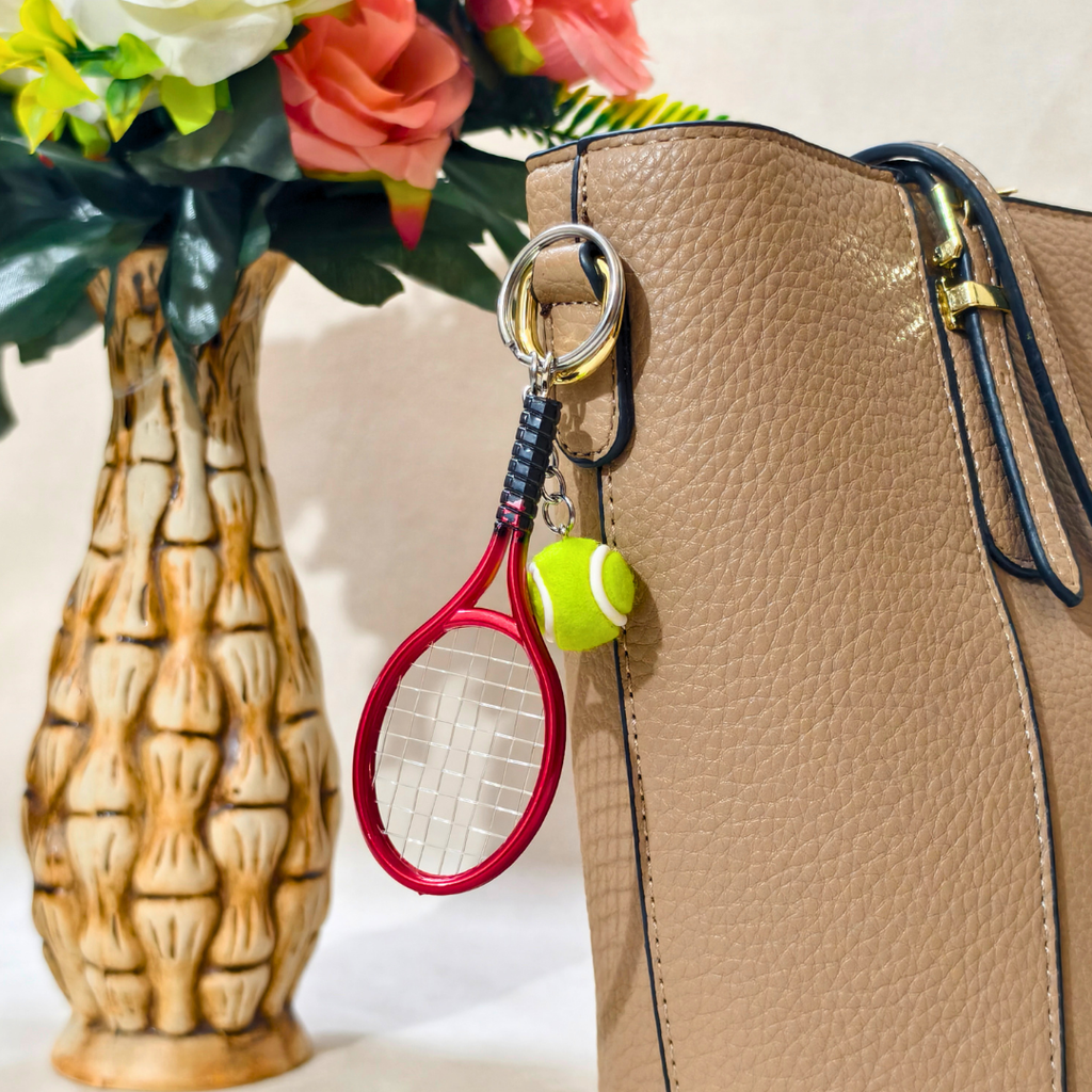 A close-up of a tan pebbled leather handbag with a red tennis racket and yellow tennis ball keychain attached near the top handle, next to a decorative floral vase.