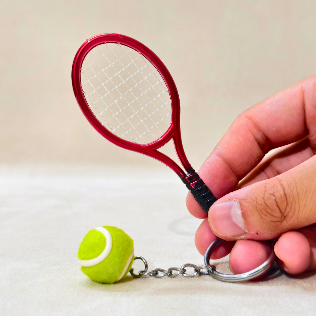 A miniature red tennis racket keychain with a small green and white tennis ball charm, held by a hand against a neutral beige background.