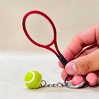 A miniature red tennis racket keychain with a small green and white tennis ball charm, held by a hand against a neutral beige background.