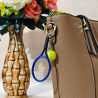 A miniature tennis racket keychain with a small yellow tennis ball charm, hang on Handbag as Bag charm against a neutral beige background.