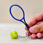 A miniature tennis racket keychain with a small yellow tennis ball charm, held by a hand against a neutral beige background.