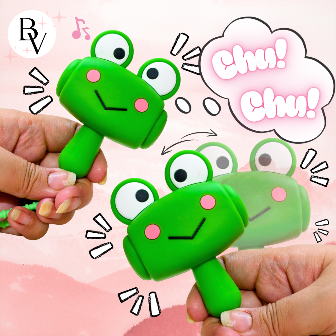 Two hands holding green frog-shaped rattle toys against a pink background with cartoon speech bubbles and decorative marks. A circular 'BV' logo appears in the top left.