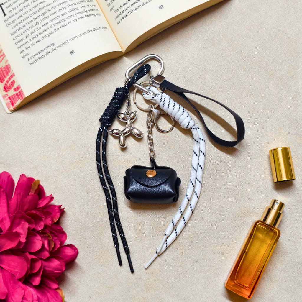 Black keychain with a small pouch and strap, next to a pink flower and an open book on a beige surface.
