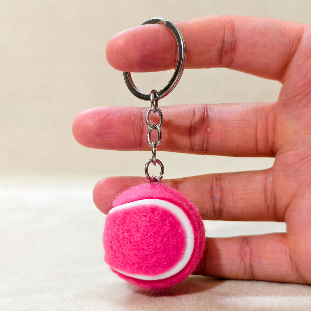 A small pink felt tennis ball keychain held between a person's fingers. The keychain features a bright pink felt ball with a white curved stripe, attached to a silver metal ring and chain.