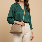 Woman wearing a green blouse and white pants, holding a beige handbag against a beige background