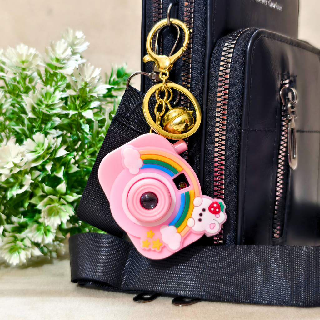 Pink camera-shaped keychain with gold accents on a black bag with greenery in the background