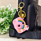 Pink camera-shaped keychain with gold accents on a black bag with greenery in the background