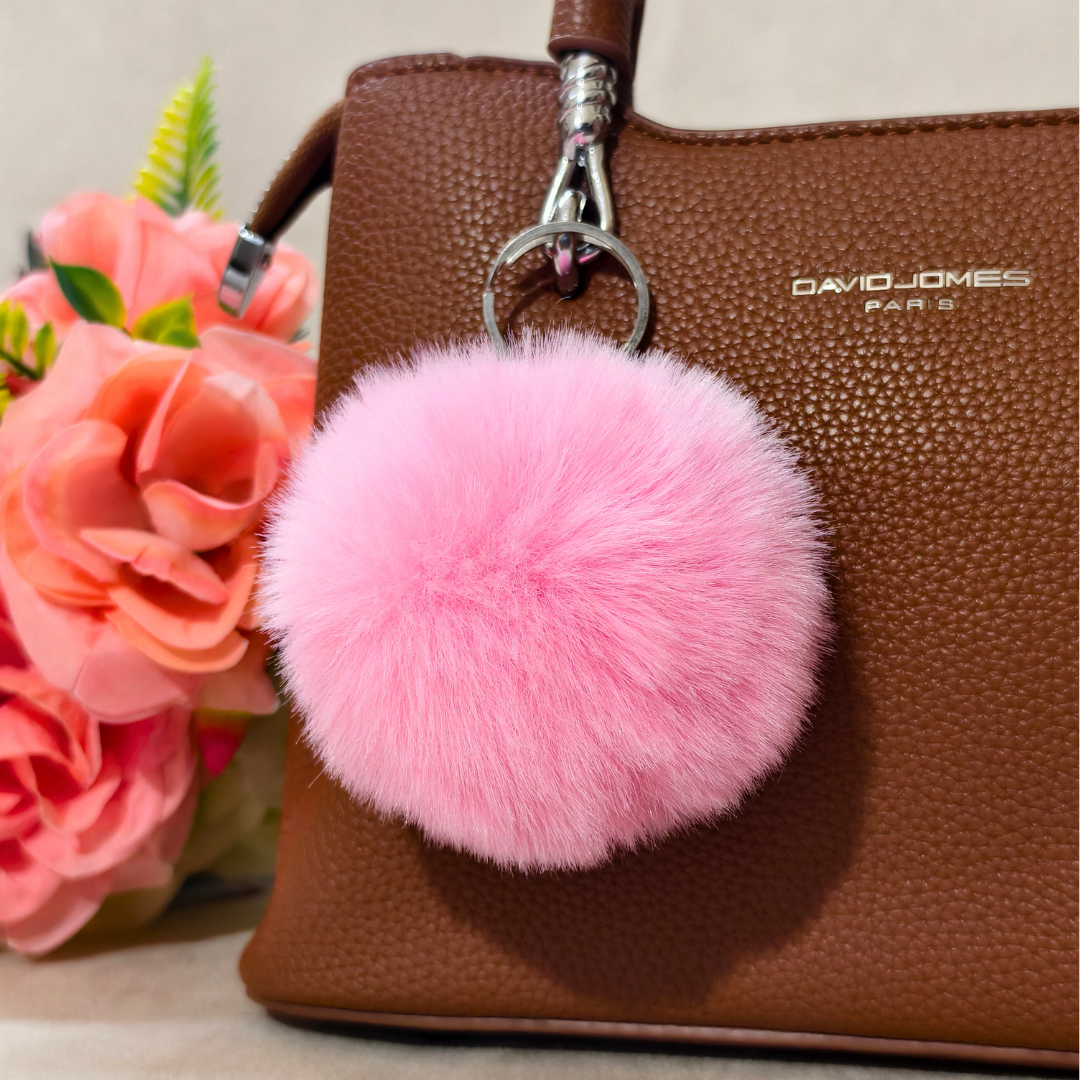 Pink fluffy pom-pom keychain on a brown handbag with flowers in the background
