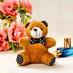 A small plush teddy bear keychain or charm in warm brown with a white snout, black polka-dot bow tie, and black polka-dot ear and paw pads, posed on a light beige surface with pink flowers and a blue perfume bottle in the background.