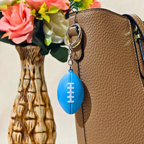 A blue football-shaped keychain with white laces hanging from a gold ring on a brown textured handbag, with a decorative vase of flowers in the background.