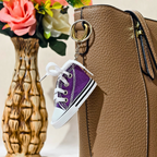 A miniature purple canvas sneaker keychain charm with white laces and a white rubber toe cap, hanging from a gold ring attached to a brown textured handbag. A decorative vase with flowers is blurred in the background.
