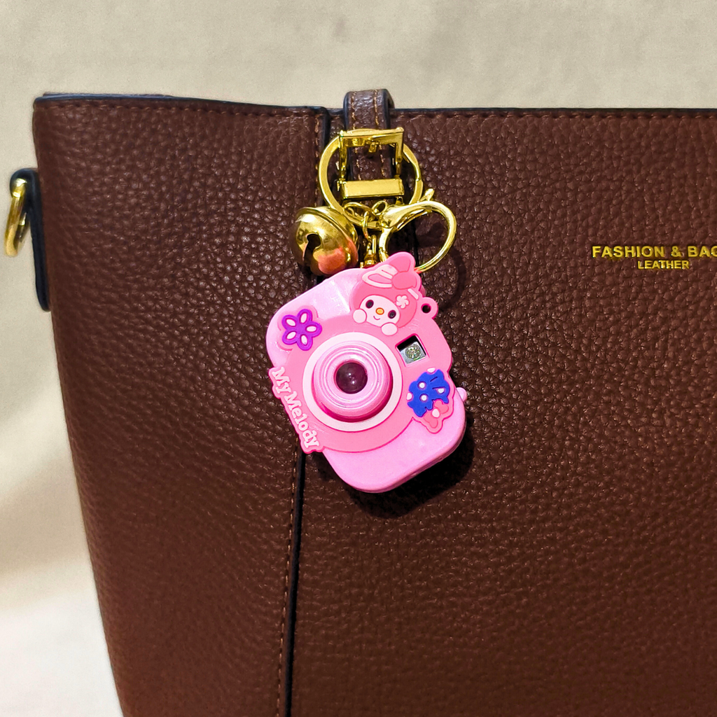 Pink camera-shaped keychain with cartoon characters attached to a brown leather bag.