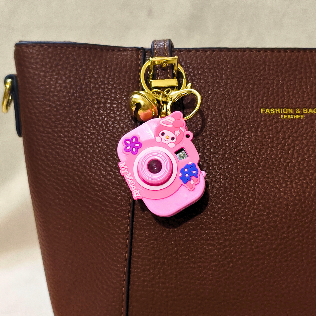 Pink camera-shaped keychain with cartoon characters attached to a brown leather bag.