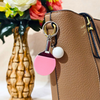 A close-up of a tan textured handbag with a pink ping-pong paddle keychain charm and a white ball accessory hanging from a gold-tone ring, with a decorative flower vase in the background.
