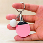 A close-up of pink ping-pong paddle keychain charm and a white ball accessory hanging from a gold-tone ring, holding in hand in the beige background.