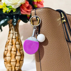 A novelty table tennis keychain featuring a miniature purple and silver paddle with a small white ball charm attached by a silver chain and key ring, Attached on Handbag as Bag Charm shown against a Beige background.