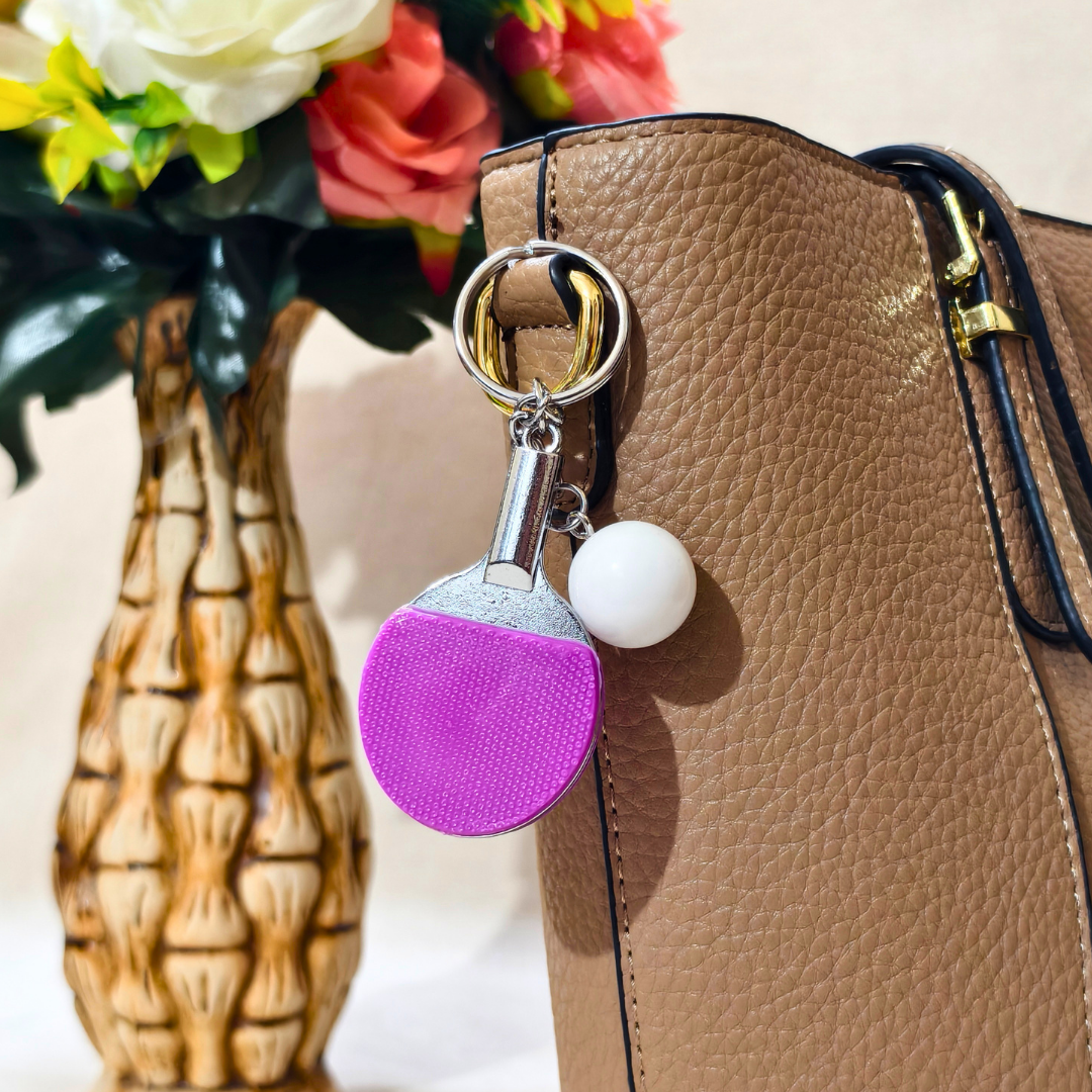 A novelty table tennis keychain featuring a miniature purple and silver paddle with a small white ball charm attached by a silver chain and key ring, Attached on Handbag as Bag Charm shown against a Beige background.