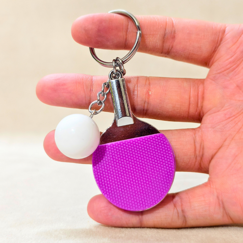 A novelty table tennis keychain featuring a miniature purple and silver paddle with a small white ball charm attached by a silver chain and key ring, hold in hand shown against a Beige background.