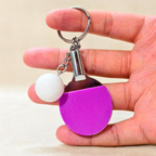 A novelty table tennis keychain featuring a miniature purple and silver paddle with a small white ball charm attached by a silver chain and key ring, hold in hand shown against a Beige background.