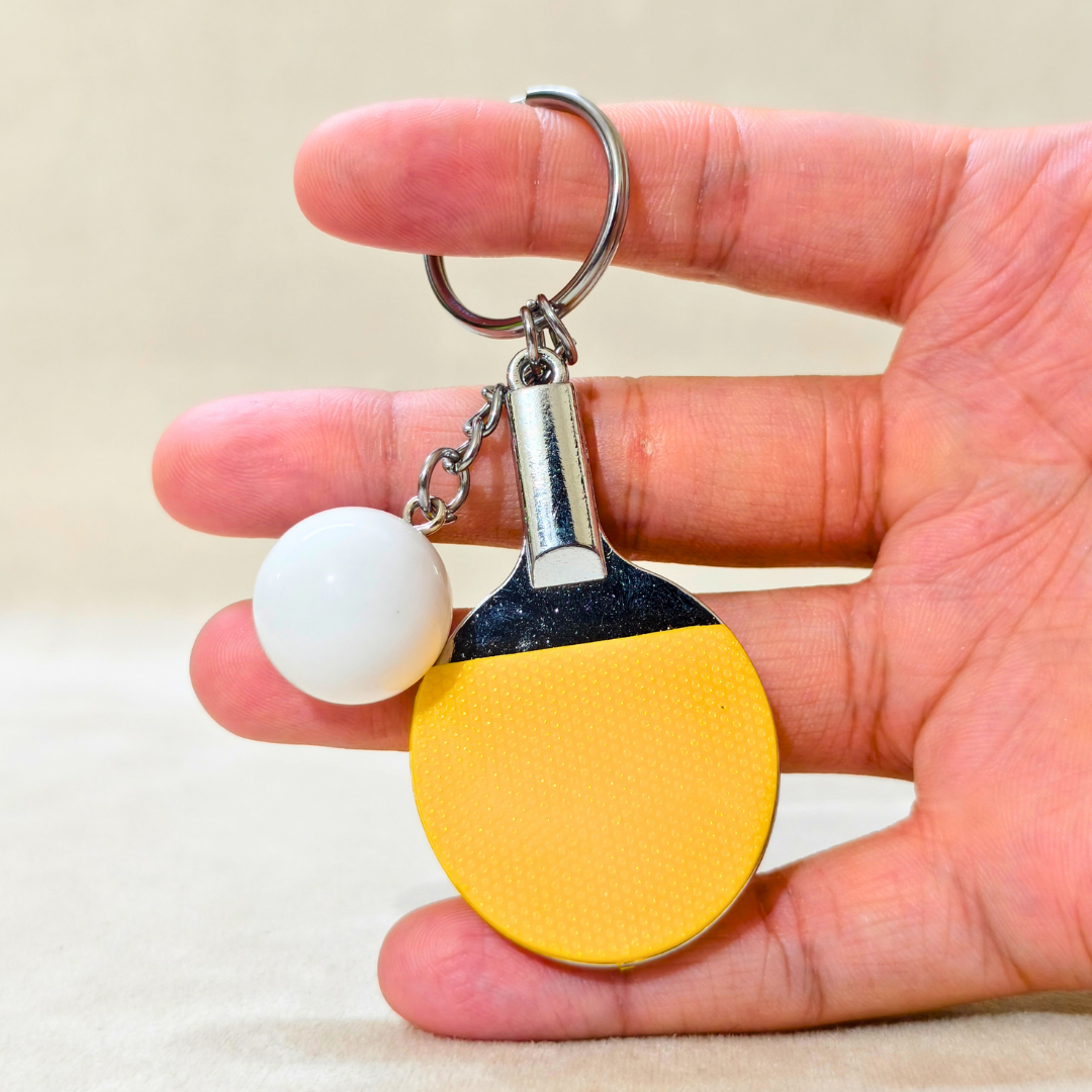 A hand holding a miniature table tennis keychain charm, featuring a yellow and black paddle with a small white ball and silver metal key ring.