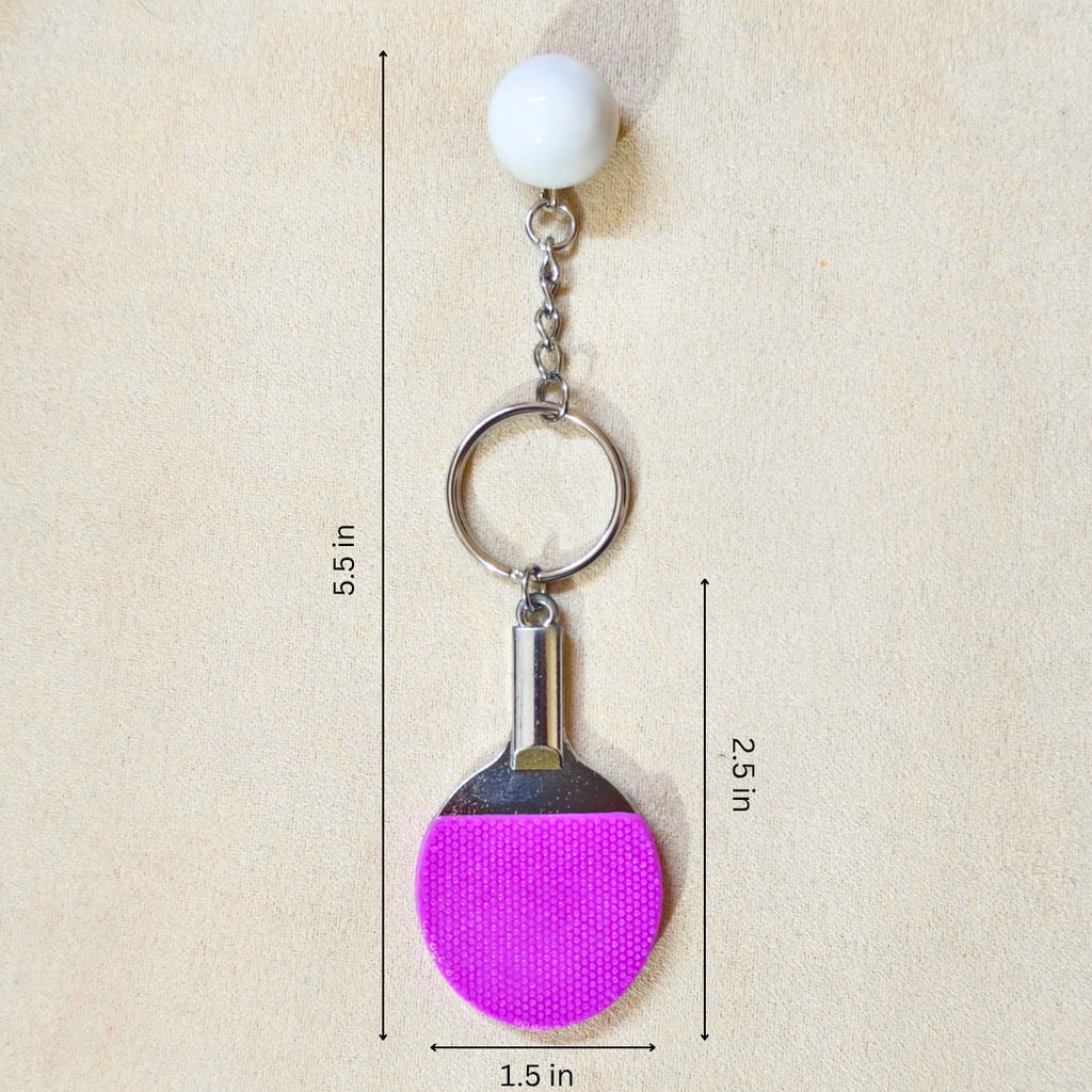 A table tennis-themed keychain with a white ball charm, silver metal chain and ring, and a pink paddle-shaped charm on a beige textured background with dimension lines and text.